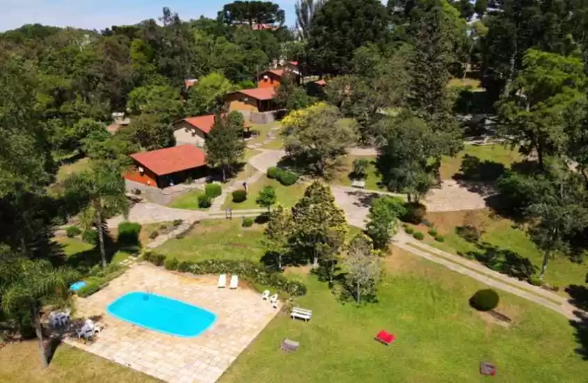 Durante um dia ensolarado, vista aérea de um hotel fazenda com bastante área verde, piscina, sofás de descanso, mesas, cadeiras, espreguiçadeiras e bancos