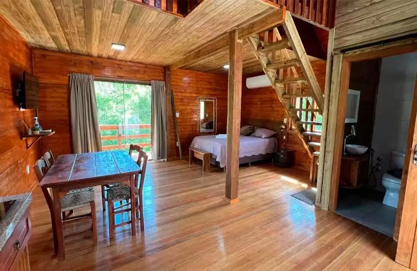 Interior de um chalé com banheiro, cama de casal, TV, janela grande, ar condicionado, mesa, cadeiras e estrutura de madeira
