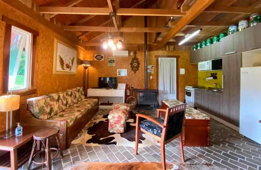 Sala de um chalé com estrutura e móveis de madeira, sofás, cadeira, banco, tapete, TV, quadros decorativos e cozinha integrada