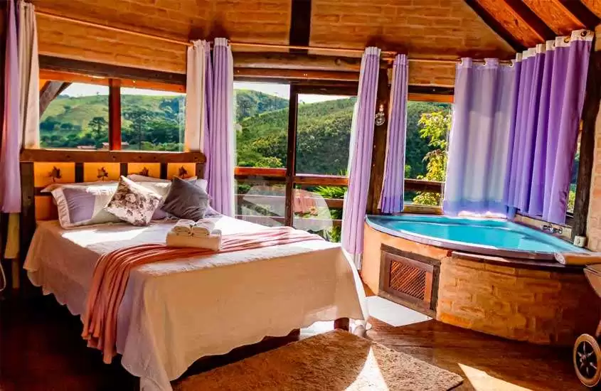 Interior de um chalé com cama de casal, banheira de hidromassagem janelas grandes acortinadas com vista das montanhas e móveis de madeira