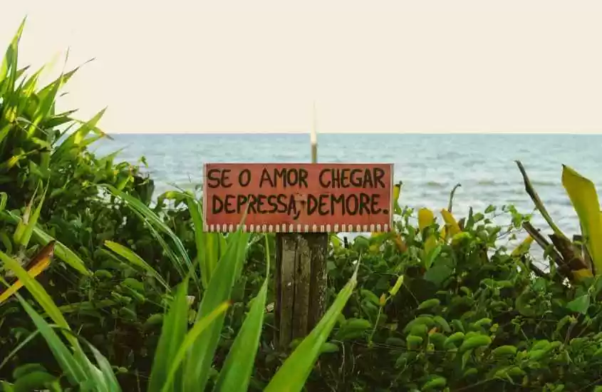 Durante o entardecer praia de Caraíva com vegetação verdinha, mar no fundo e placa escrito "se o amor chegar depressa, demore."