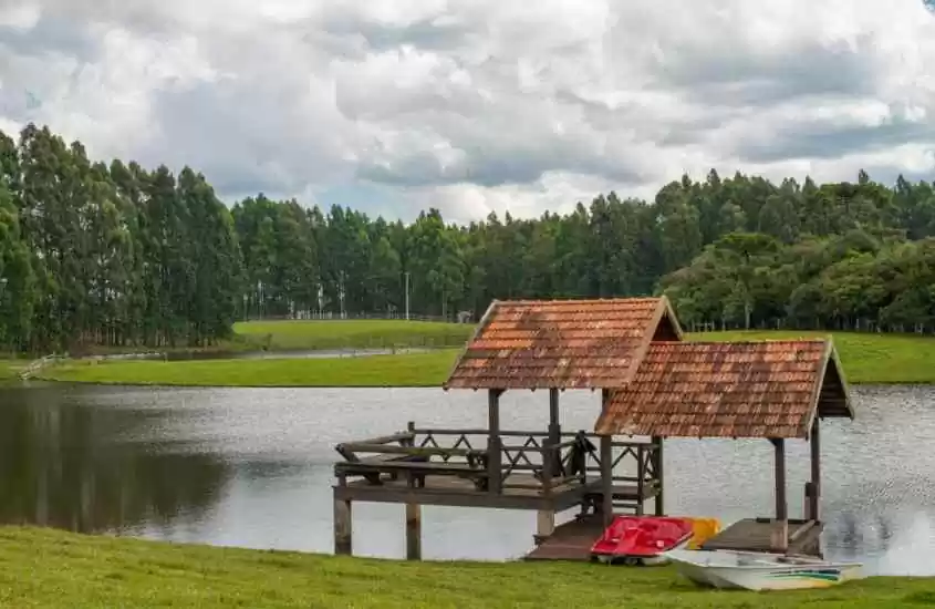 Em um dia nublado, lago de um hotel fazenda no rio grande do sul com decks de madeira cobertos, pedalinhos, barco e árvores ao redor