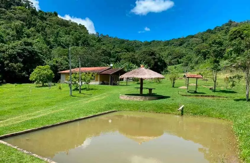 Em um dia de sol, área de lazer de Hotel Fazenda na Serra da Mantiqueira com lago, coreto, banco, árvores e serra ao redor