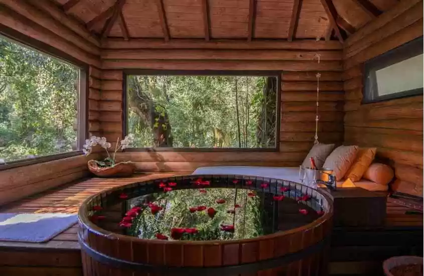Banheira de um dos hotéis fazenda no rs com deck de madeira, cama atrás, janelas grandes ao redor, flores e balde de champagne com taças