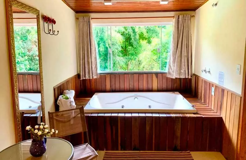 Área de banheira com deck de madeira, cadeira, mesa, espelho e janela grande acortinada