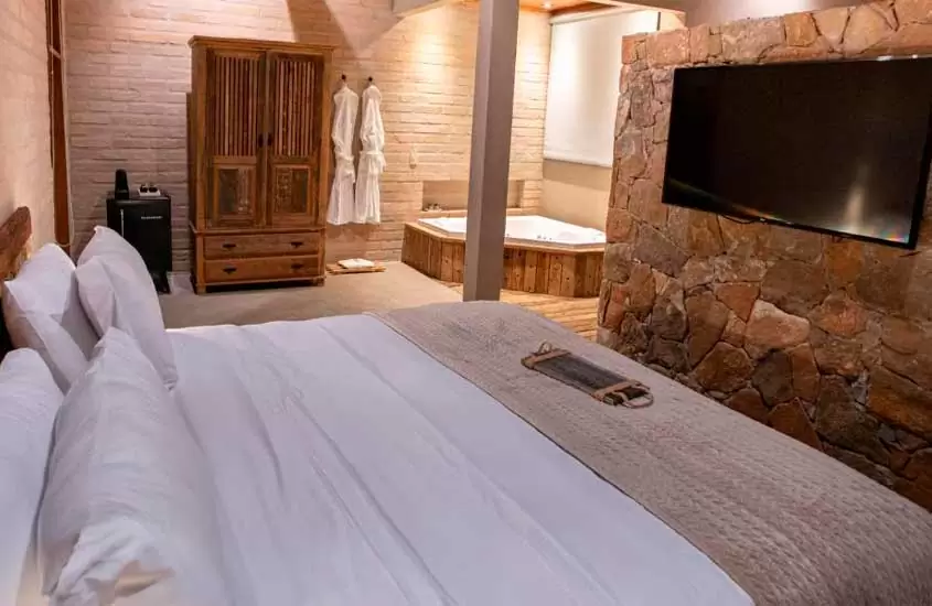 Quarto de um hotel com cama de casal, TV, parede de pedra, armário de madeira, frigobar, roupões e banheira
