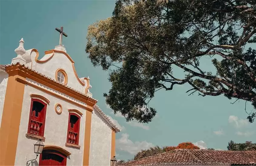 Em um dia de sol, vista da fachada de uma igreja em Tiradentes com janelas e portas vermelhas, e árvores ao redor