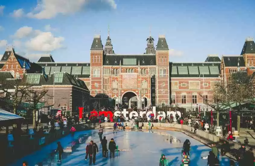 Em um dia de sol, pista de patinação no gelo em Amsterdam com pessoas, árvores secas, letreiro escrito "I amsterdam" e prédio atras