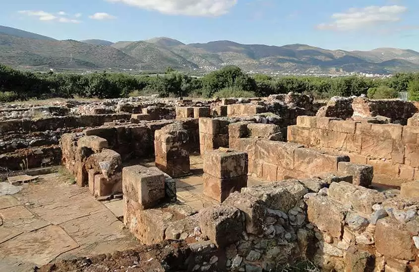 Em um dia de sol, uma ótima opção de o que fazer em Creta é a visita das ruinas do Palácio de Malia, com diversas árvores ao redor e montanhas de fundo