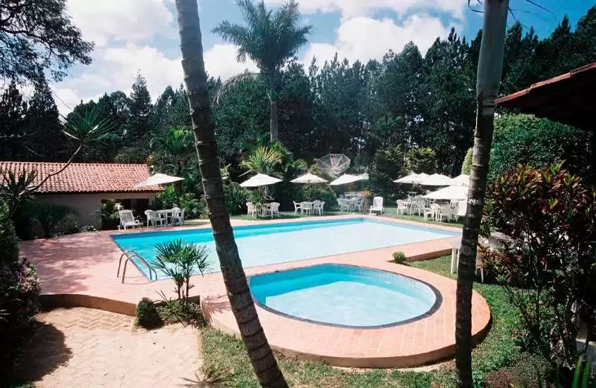 Durante um dia ensolarado, área de lazer de um hotel fazenda perto de juiz de fora com piscinas, mesas, cadeiras, guarda-sóis, árvores e parte gramada ao redor