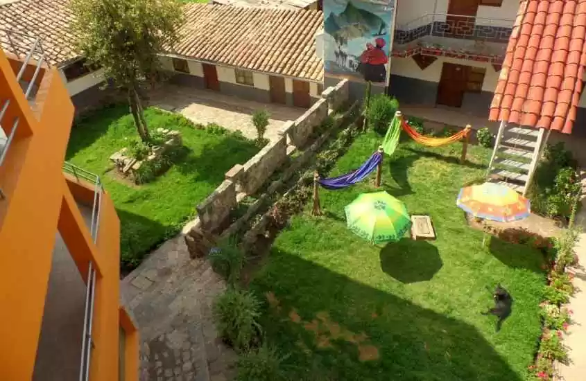 Em um dia de sol, área externa de hostel em Cusco com guarda-sóis, redes, parte gramada, árvores e plantas