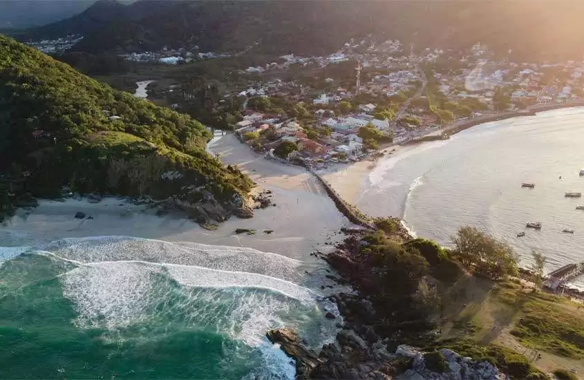 Em um final de tarde, paisagem aérea de praia em Florianópolis, lugar onde ir no feriado de corpus christi, com praia, árvores ao redor e cidade no fundo