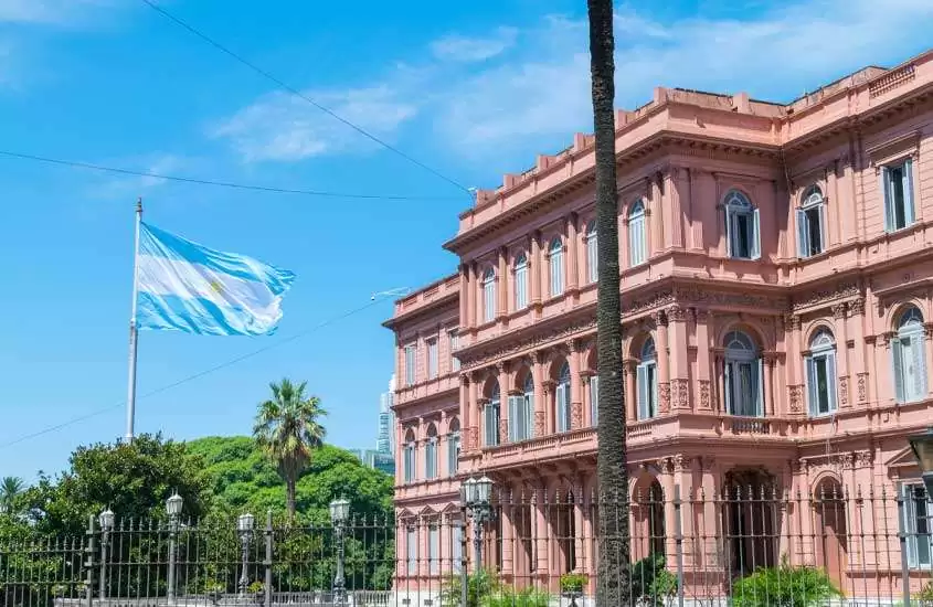 Durante uma manhã de sol, fachada de um monumento de Buenos Aires com bandeira hasteada na frente, grade e árvores ao redor
