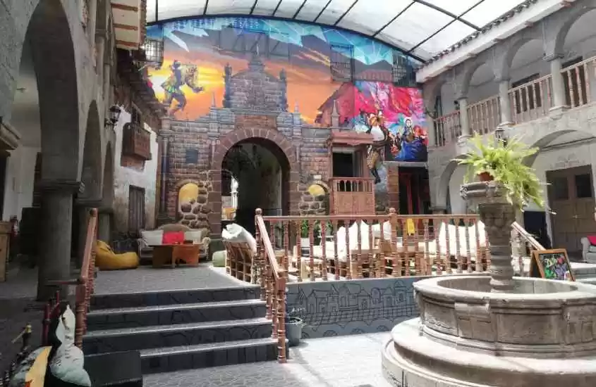 Área externa de um hostel em Cusco com mesas, cadeiras, sofás, puffs, chafariz e parede com pinturas temáticas