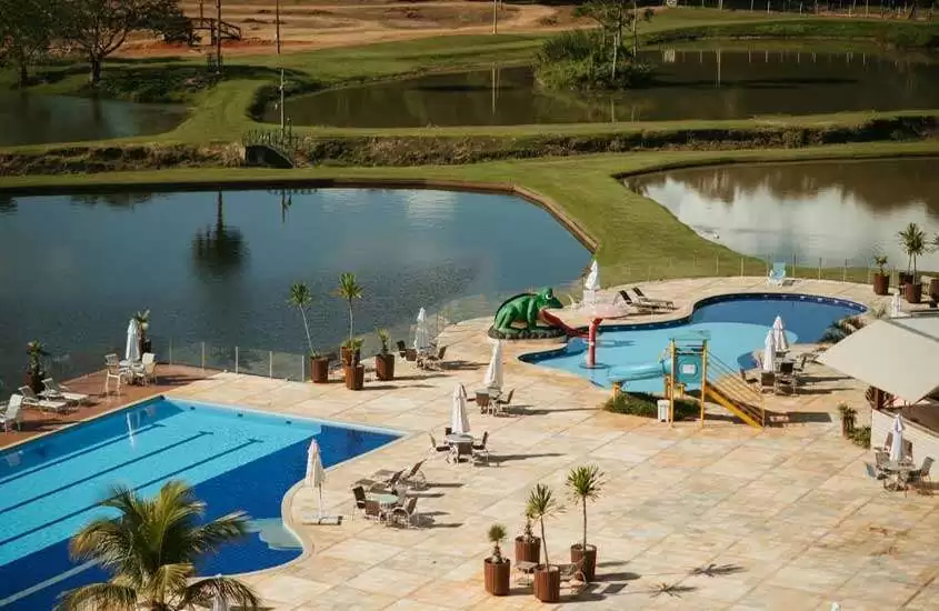 Em um dia de sol, área de lazer de um resort com lagos, piscinas, espreguiçadeiras, mesas, cadeiras, guarda-sóis, escorregadores infantis, árvores e parte gramada ao redor