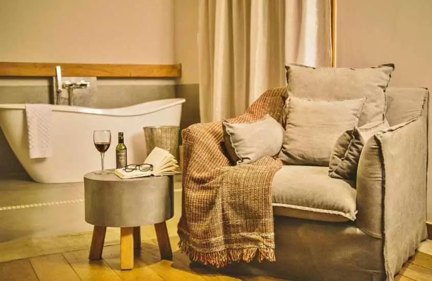Interior de quarto com poltrona, mesa, vinho, óculos, livro, poltrona com almofadas e manta, banheira com toalha e janela acortinada