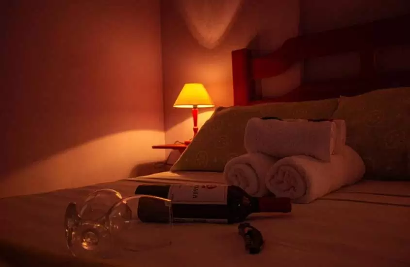 Quarto de pousada com luminária de luz amarela, cama de casal, toalhas, quadro decorativo, vinho e taças