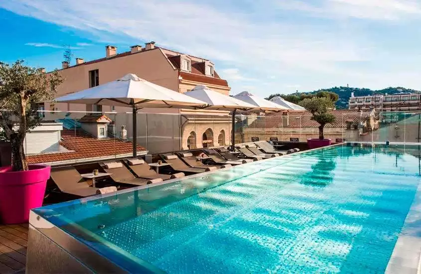 Em um dia de sol, área de lazer de um hotel onde ficar em Cannes com piscina de borda infinita, espreguiçadeiras, guarda-sóis e plantas ao redor