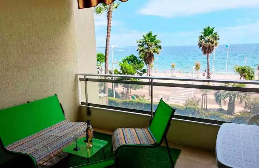 Varanda de um hotel onde ficar em Cannes com sofá, cadeiras, bebida alcoólica, árvores ao redor e praia na frente
