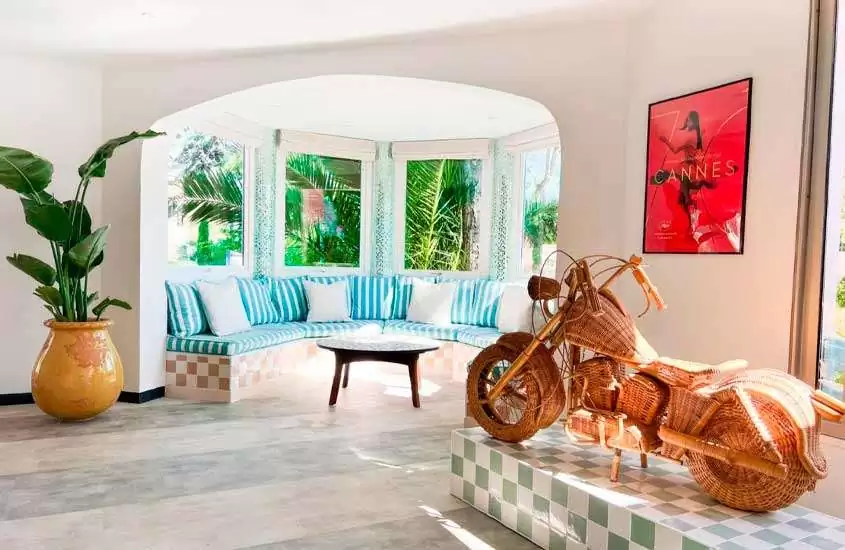 Sala de hotel com escultura, de moto, vaso de planta decorativo, canto alemão, mesa, quadro e janela grande com vista para a natureza