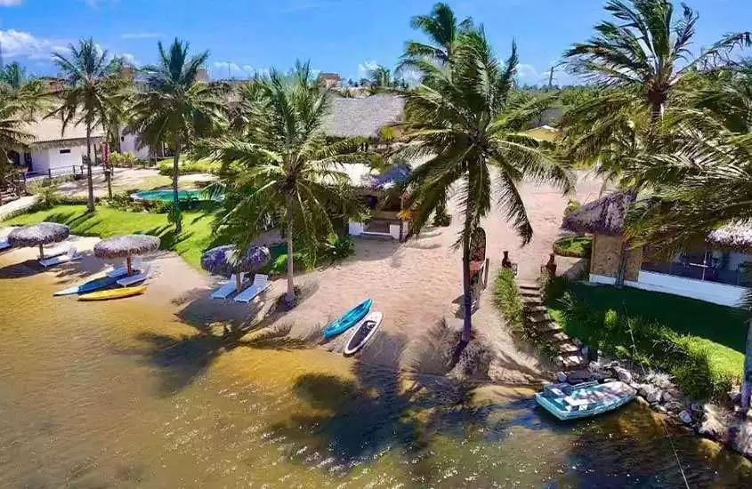 Em um dia de sol, vista aérea de praia com árvores, barcos, tendas de palha e parte gramada