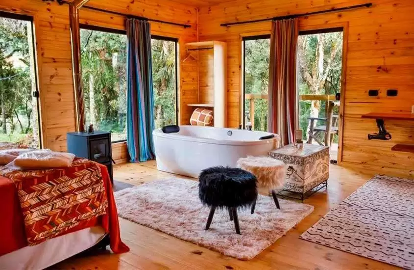 Interior de cabana na serra catarinense com cama de casal, tapetes, banheira e janelas grandes acortinadas