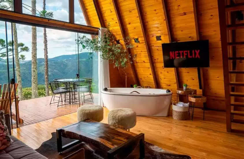 Interior de uma das cabanas na Serra Catarinense com mesas, cadeiras, sofá, banheira, TV, varanda, deck de madeira e paisagem da serra