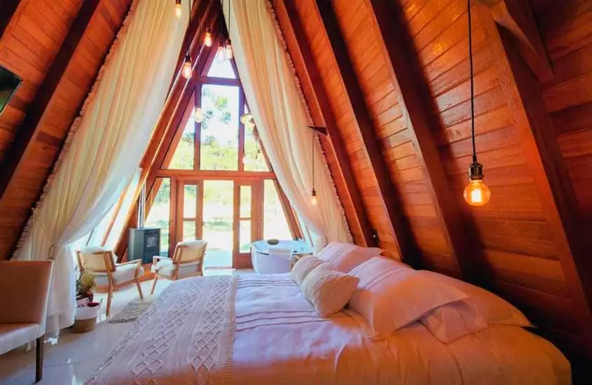 Interior de cabana de madeira com cama de casal, banheira de hidromassagem, cadeiras e cortinas