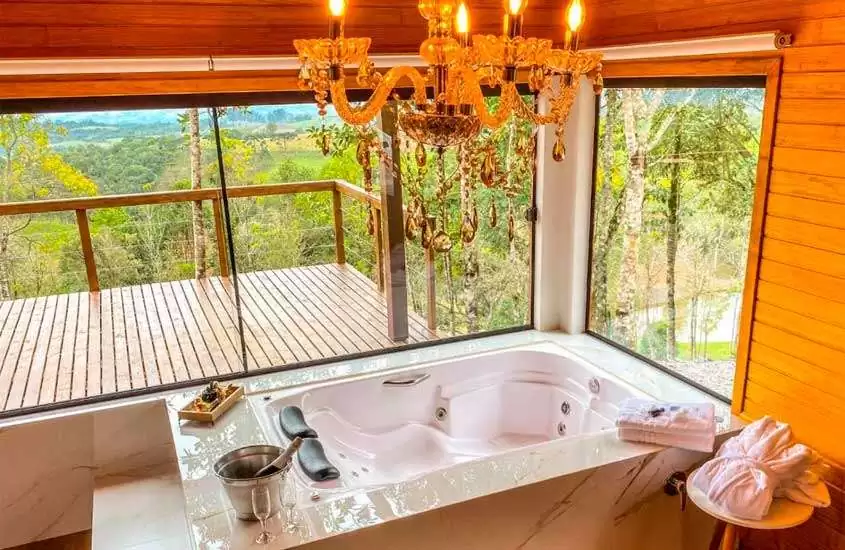 Interior de uma cabana com banheira de hidromassagem, toalhas, balde de champagne e varanda com deck de madeira e paisagem da serra