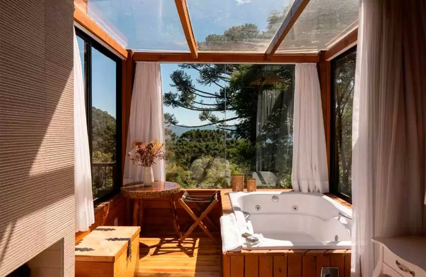 Banheira de hidromassagem de cabana na serra catarinense com deck de madeira,, bau, banco e mesa de madeira, lareira e janelas grandes acortinadas