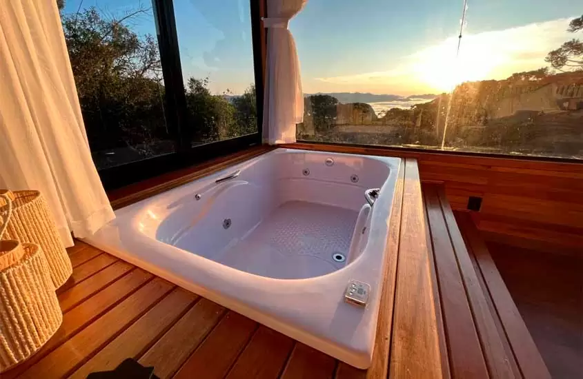 Deck de madeira com banheira de hidromassagem, janelas grandes acortinadas com paisagem da serra e decoração de cizal