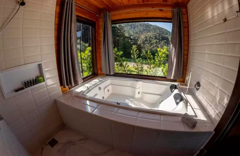 Banheira de hidromassagem de chalé na serra catarinense com chuveiro, janela acortinada com paisagem da serra