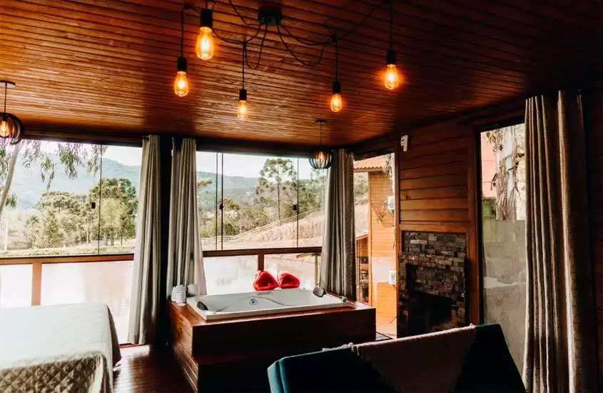 Interior de cabana na serra catarinense com cama de casal, banheira de hidromassagem, lareira e janela grande acortinada