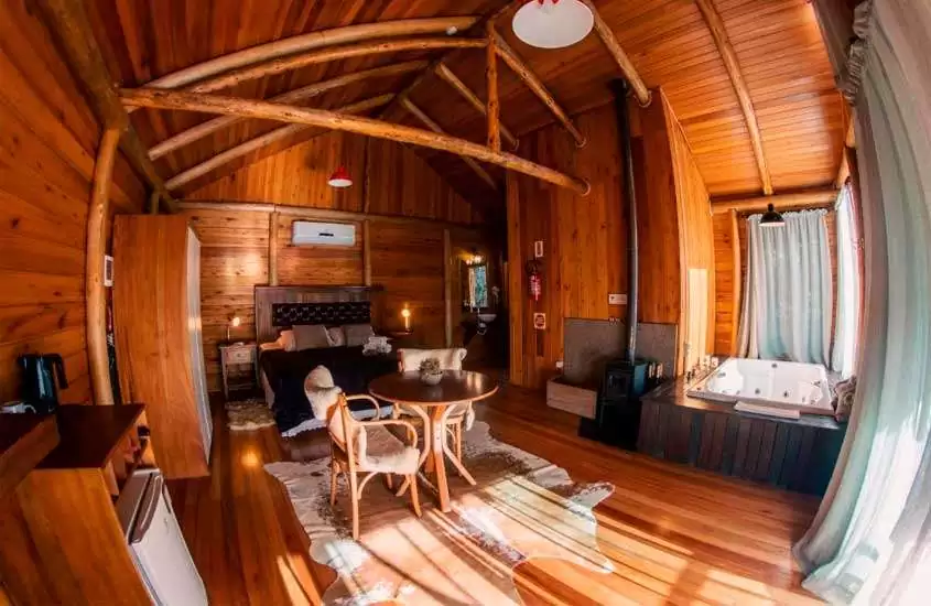 Interior de uma cabana com mesa, cadeiras, tapetes, banheira de hidromassagem, cama de casal, frigobar, lareira e ar condicionado