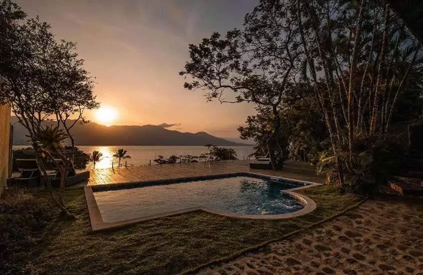 Durante o pôr do sol, área de lazer de hotel em ilhabela com piscina, mar no fundo, espreguiçadeiras, deck de madeira e árvores ao redor