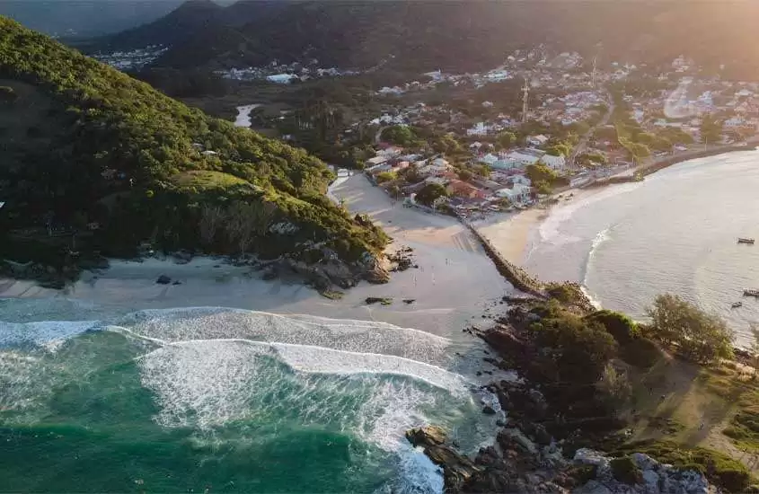 Em um dia ensolarado, vista aérea de Floripa com mar do lado, paredões de pdra cobertos de árvores e cidade no fundo