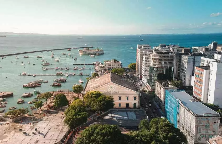 Em um dia de sol, vista aérea de Salvador com prédios, mercado, árvores ao redor, barcos aportados e praia atrás