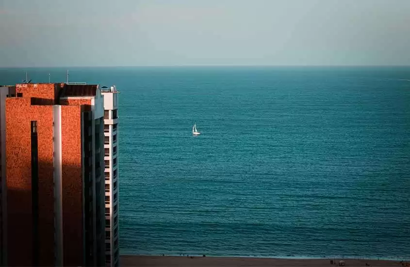 Durante um final de tarde, paisagem de uma praia onde passar o ano novo no nordeste com prédio do lado e barco no meio do mar